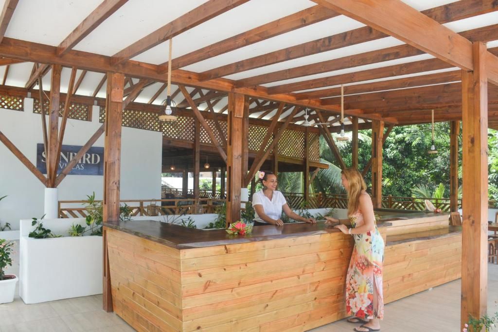 A wooden reception desk with a sign above it, surrounded by plants and a wooden pergola structure.