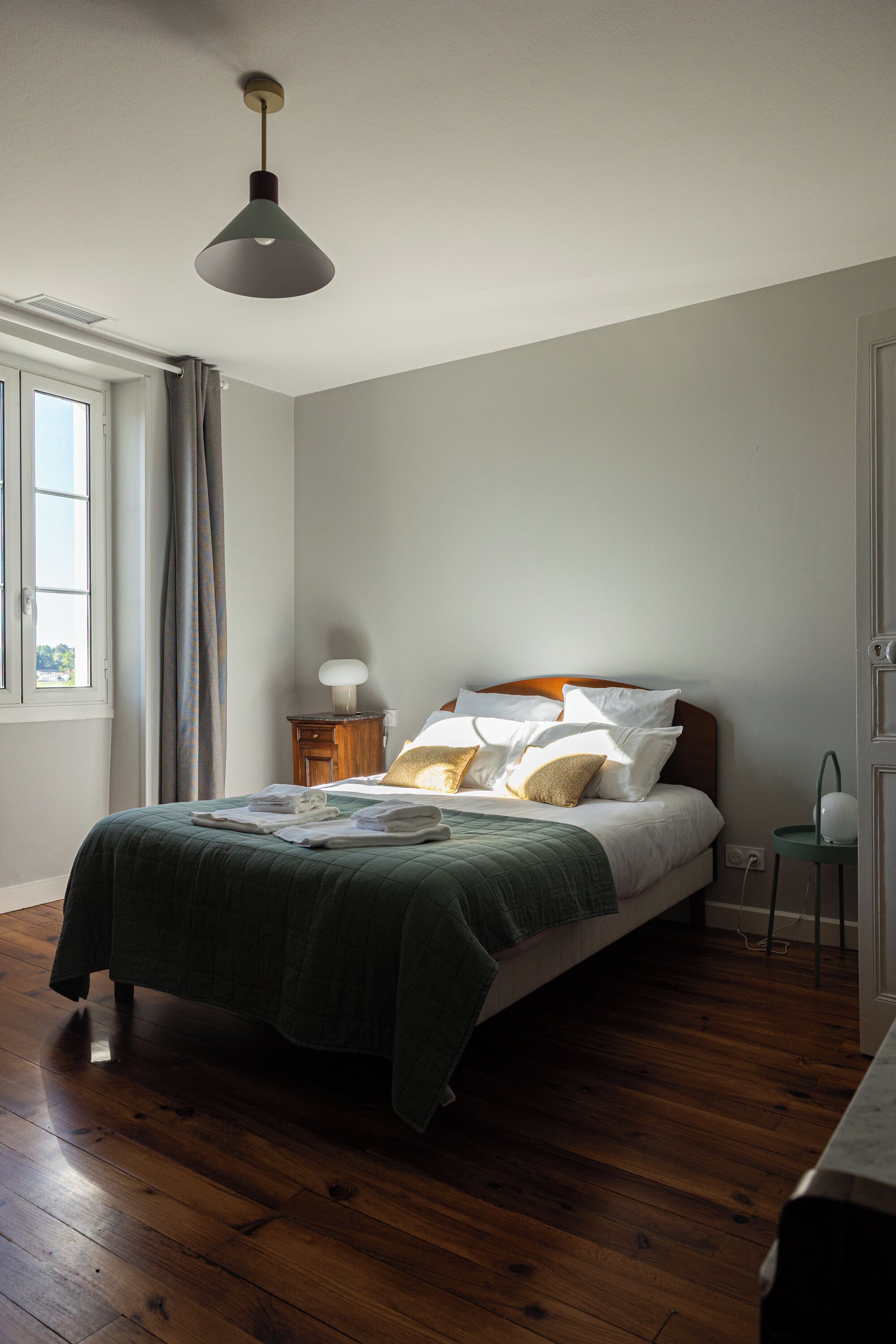 A bedroom with a bed, a bedside table, a lamp, a window with curtains, and a wooden floor.