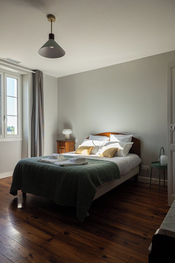 A bedroom with a bed, a bedside table, a lamp, a window with curtains, and a wooden floor.
