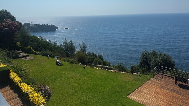 Paesaggio costiero con area verde, terrazza in legno e vista sul mare.
