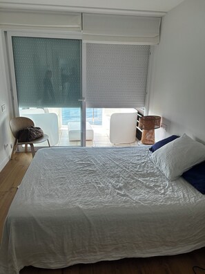 Ein Schlafzimmer mit einem großen Bett, einem Sessel, einem kleinen Tisch und Meerblick durch eine Schiebetür.