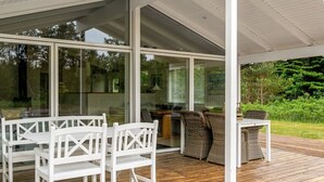 Área externa coberta com deck de madeira, móveis brancos e portas de vidro que dão acesso a um espaço interno.