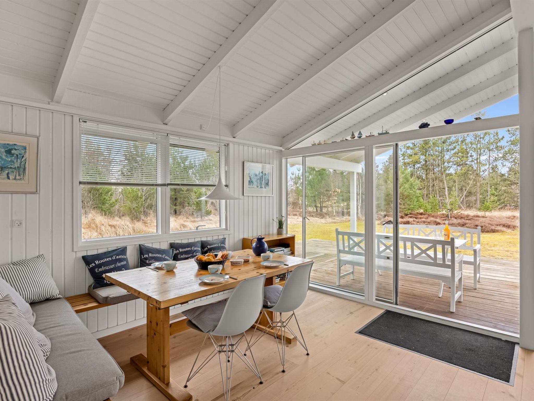 A spacious room with a wooden dining table, white chairs, and a bench. There are large windows with blinds, a ceiling fan, and a view of the outdoors.