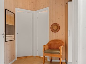 Une pièce d’angle avec deux portes blanches, une chaise en osier avec un coussin, un tableau encadré sur le mur de gauche et un chapeau de paille sur le mur de droite.