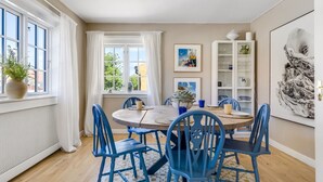Un comedor con una mesa redonda, sillas azules, un aparador blanco con cristalería y una obra de arte grande enmarcada en la pared.