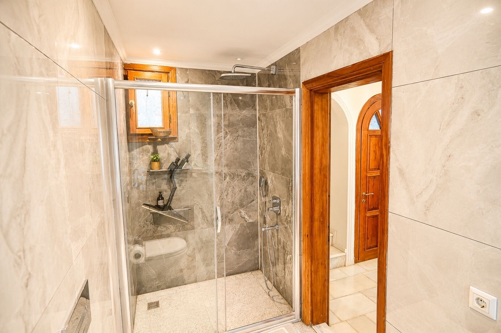 A modern bathroom with a glass-enclosed shower, marble walls, and a wooden door.