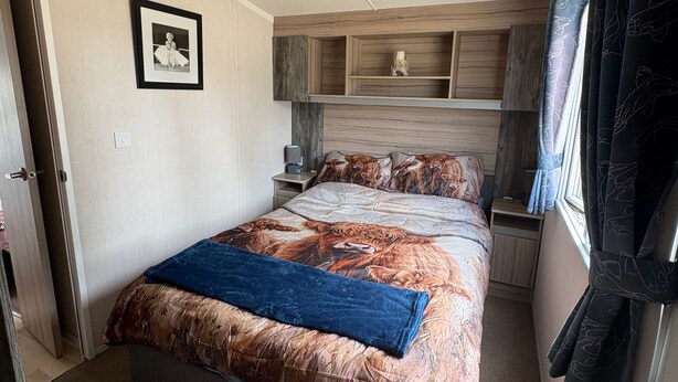 A bedroom with a bed, a bedside table, a nightstand, a picture on the wall, and a window with curtains. - Billing Aquadrome 2-bed Caravan, Sleeps 6, Parking (Northampton)