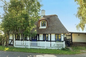 Una casa tradicional amb teulada de palla, valla blanca de palets, un petit porxo i el número '27' a la paret.