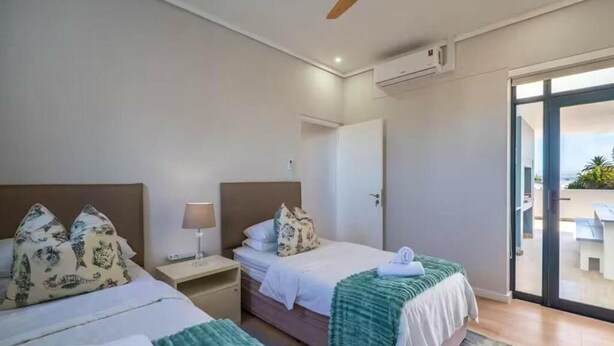 A hotel room with two beds, a ceiling fan, an air conditioning unit, a nightstand, and a view of the outside. - Kabeljauws Beachfront Luxury Apartments (Jeffreys Bay)