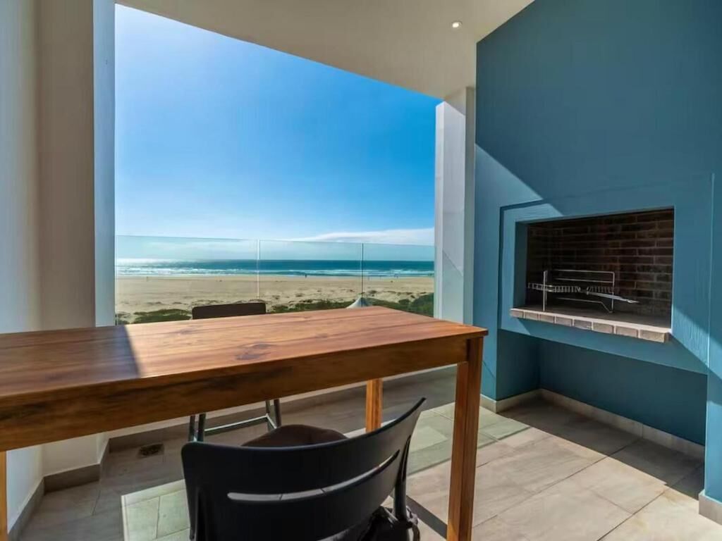 Un balcón con vista a la playa, una mesa de madera, una silla negra y una parrilla empotrada.