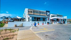 A modern building with a 'OK urban' sign, a covered outdoor seating area, and a parking lot with a handicapped accessibility symbol. - Kabeljauws Beachfront Luxury Apartments (Jeffreys Bay)