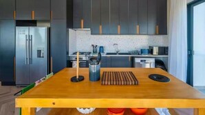 A modern kitchen with dark cabinets, a stainless steel refrigerator, a wooden table, and a window with curtains. - Kabeljauws Beachfront Luxury Apartments (Jeffreys Bay)