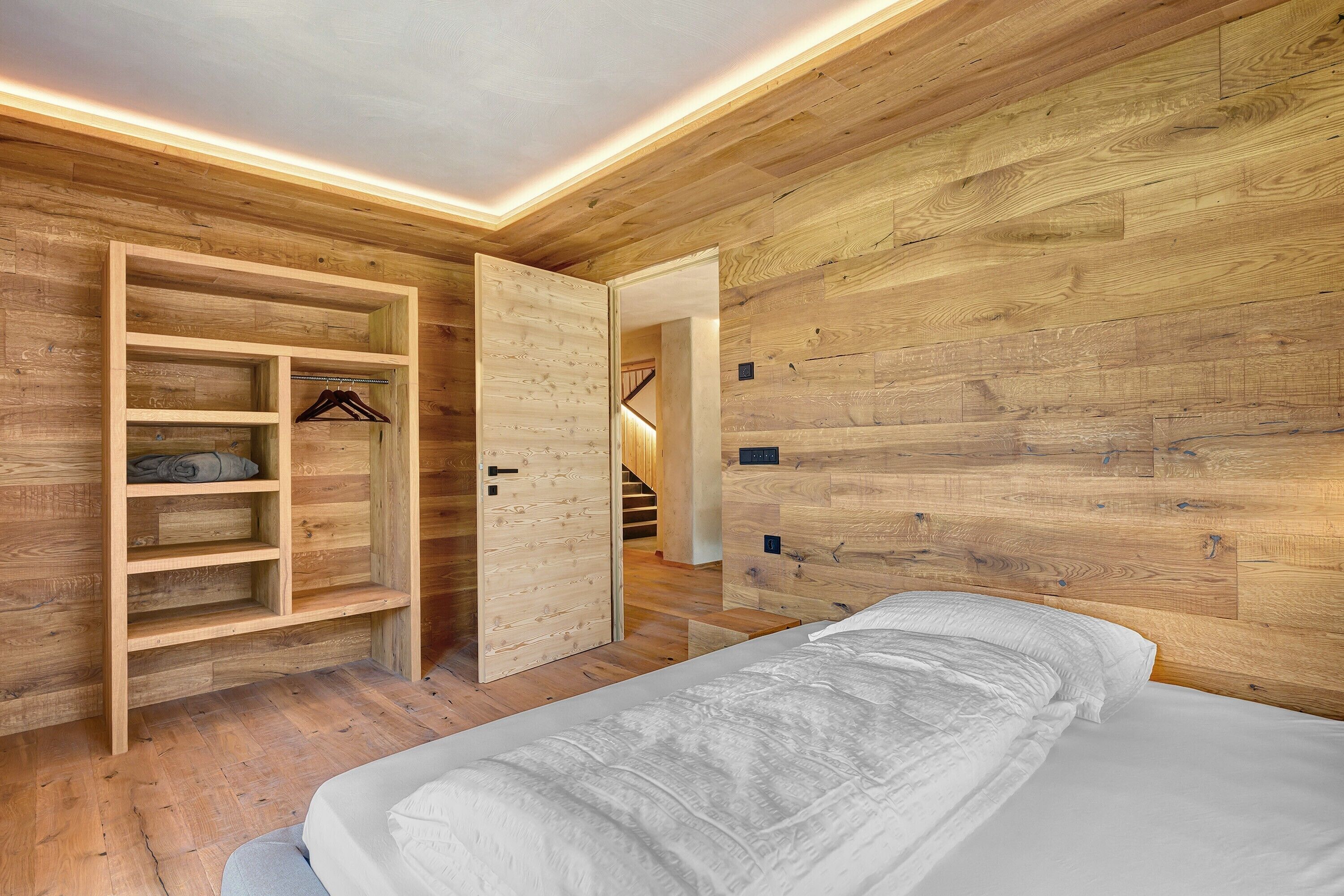 A bedroom with a bed, wooden walls, a built-in wardrobe with shelves, and a staircase leading to another level.