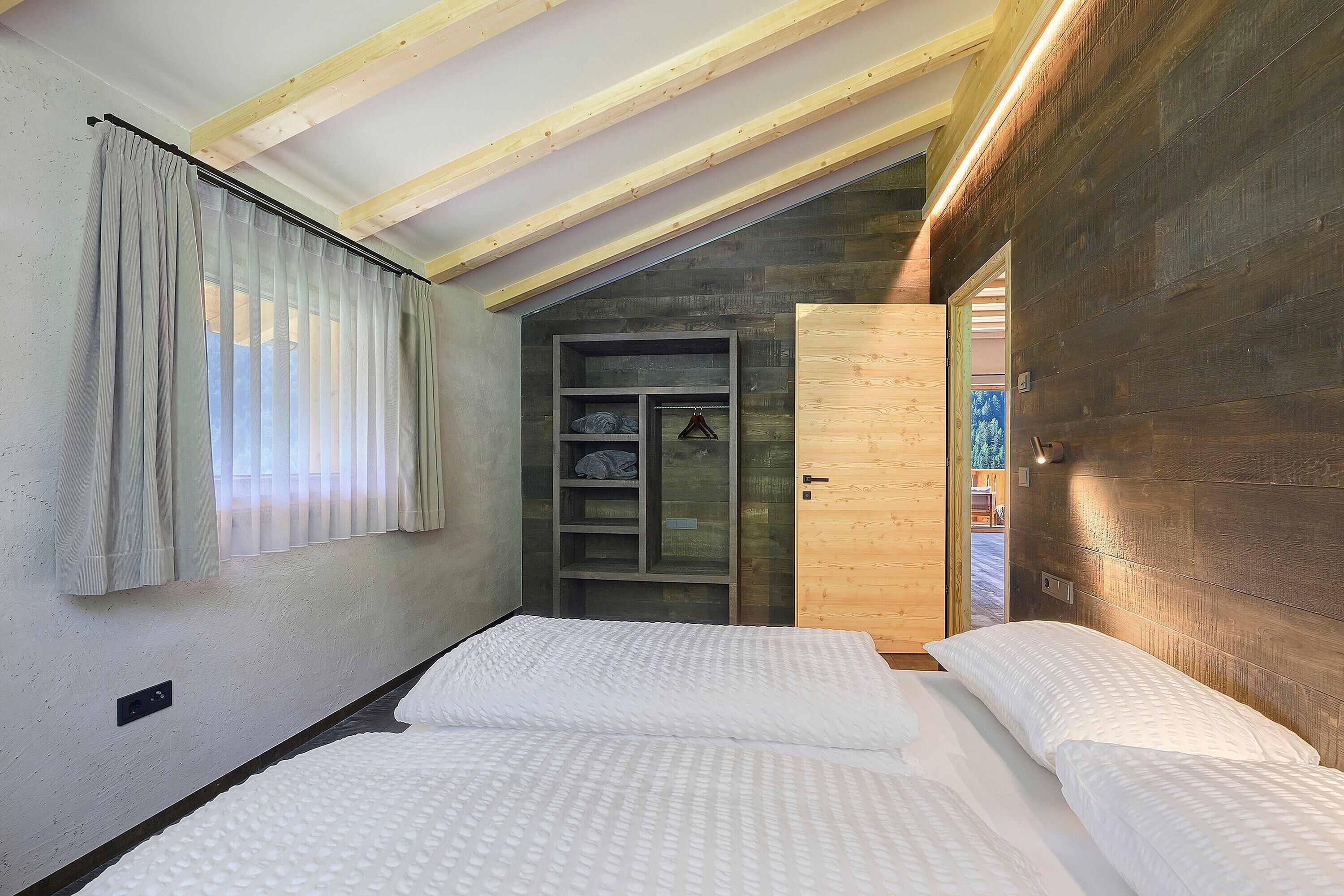 A bedroom with a sloped ceiling, a bed with white bedding, a wooden door, and a window with sheer curtains.