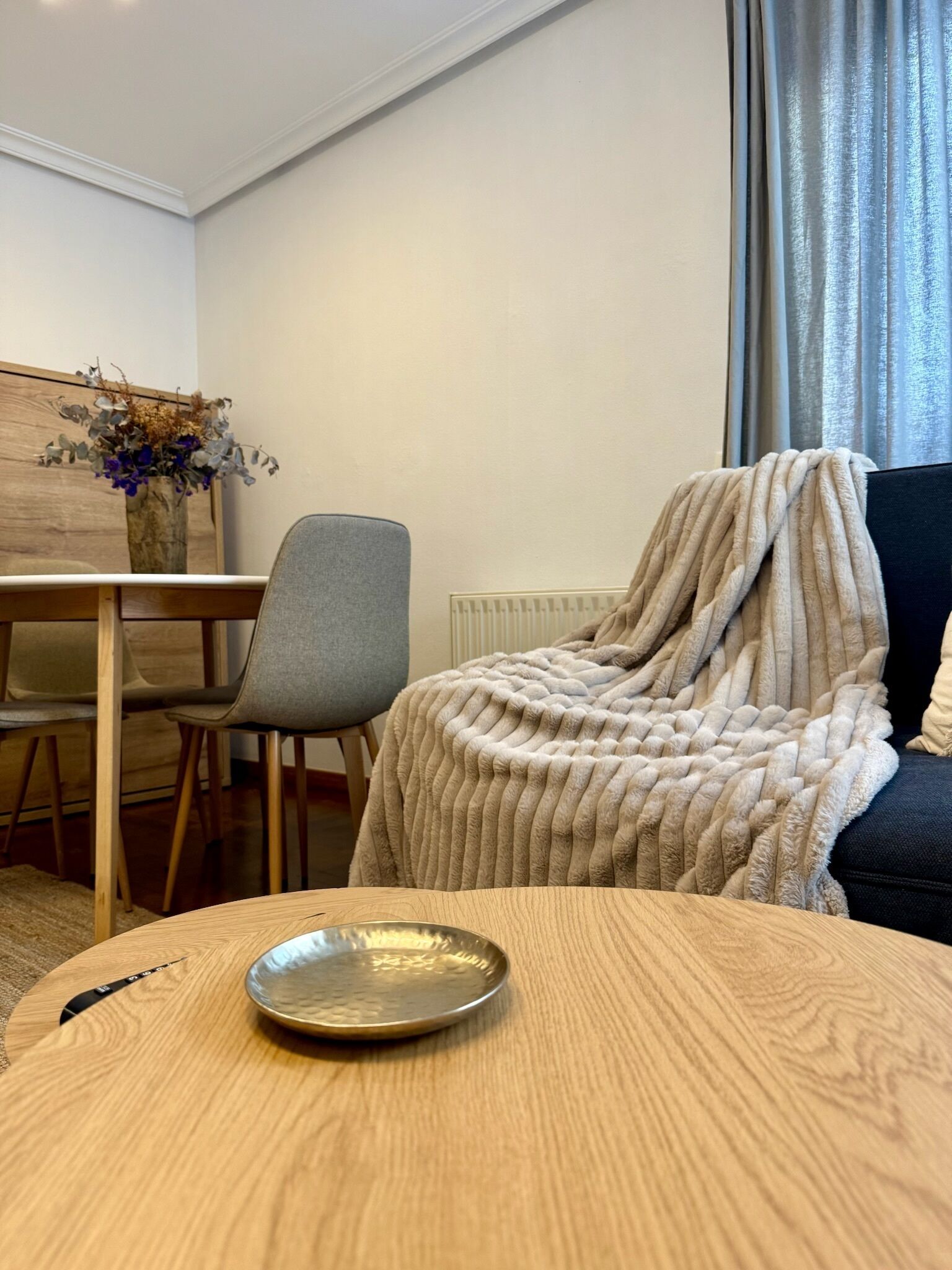 Una habitación con una mesa redonda de madera, una silla gris, un sofá con una manta, un radiador y una pared de madera con un florero.