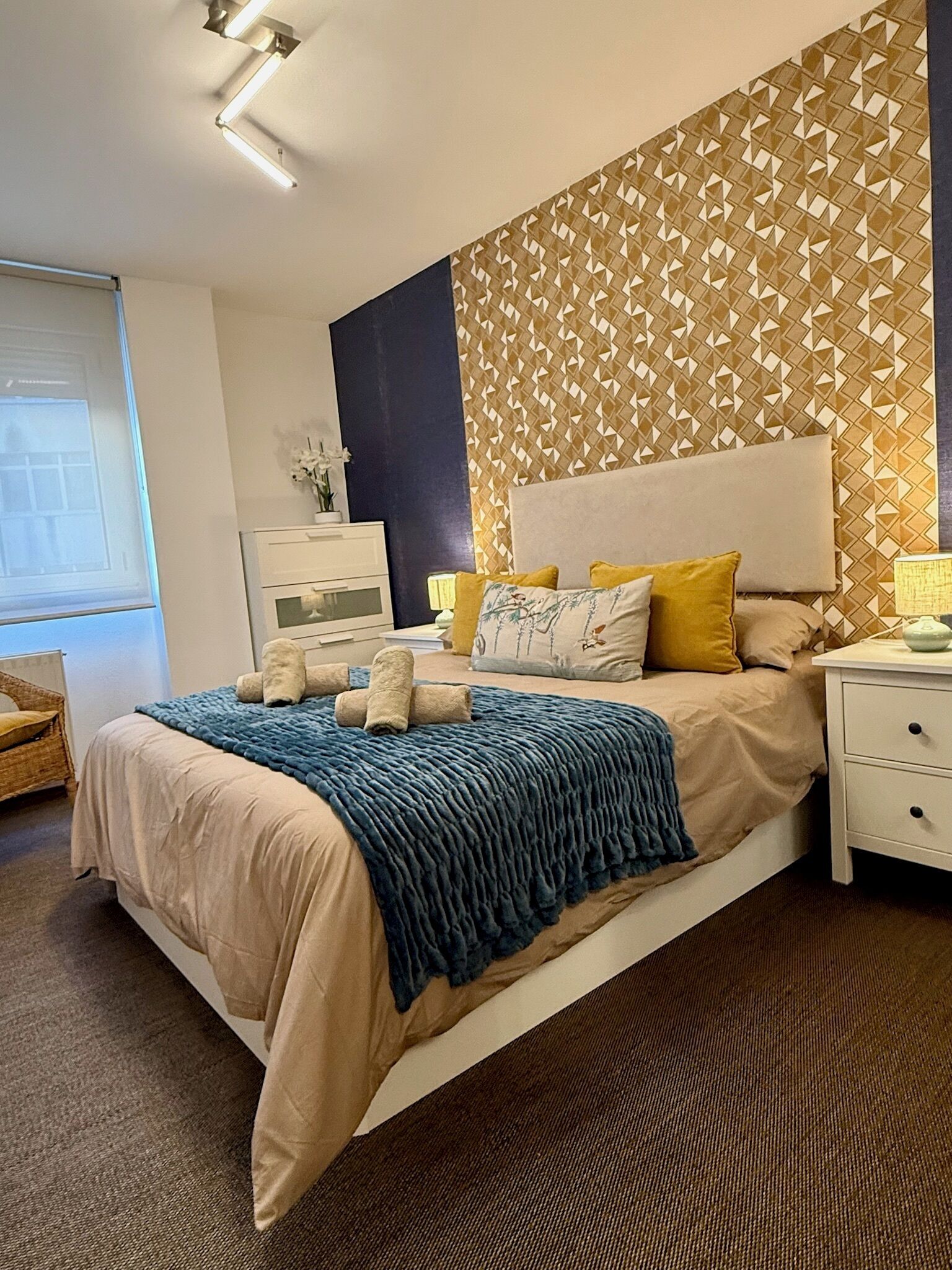 Une chambre d’hôtel comprenant un lit, des tables de chevet, une commode, une lampe, une fenêtre et un mur orné d’un motif géométrique.
