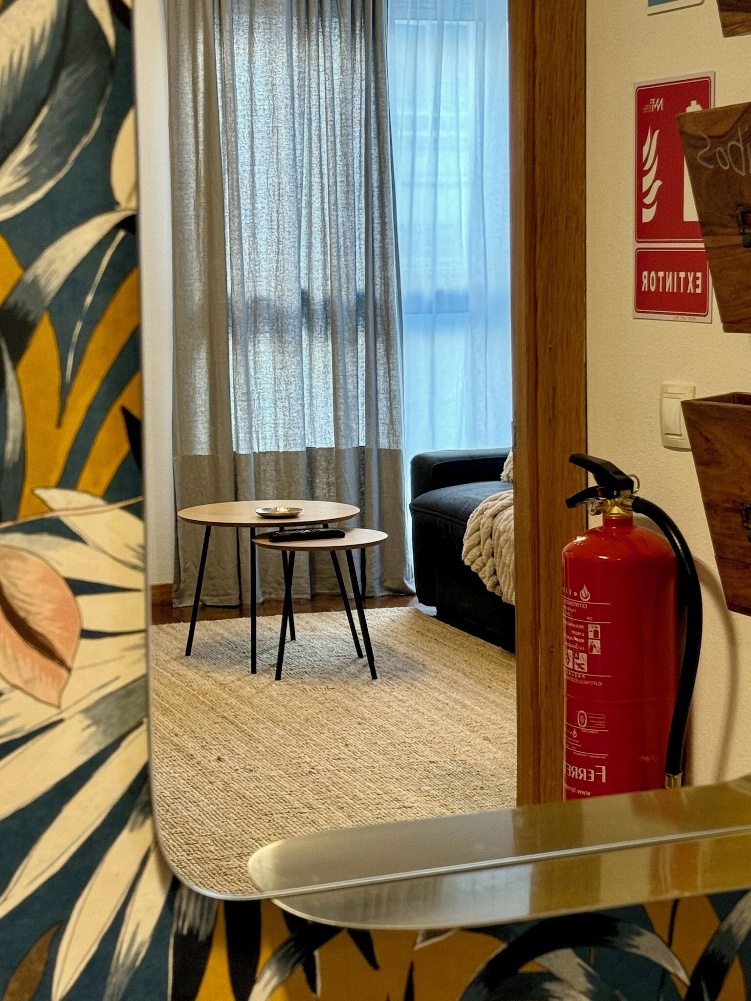 Een kamer met een kleine ronde tafel, een bank, een brandblusser en een raam met gordijnen.