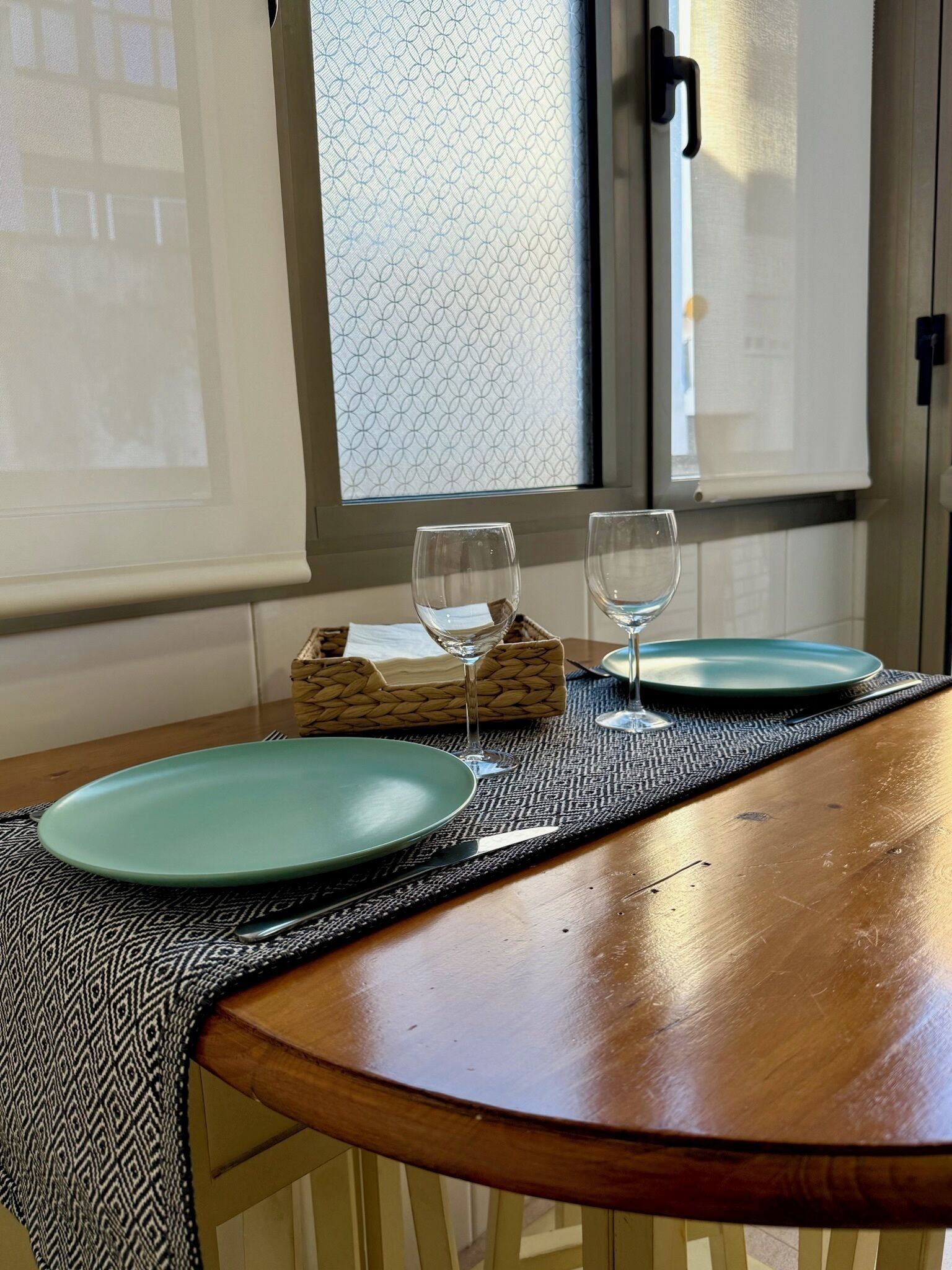 A dining table with two plates and two wine glasses, a woven basket, and a window with a geometric pattern.