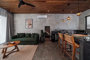 A modern living space with a green sofa, a wooden coffee table, a kitchen area with bar stools, and a ceiling fan. - Unique and Stylish Villa w/ Plunge Pool Near beach (Canggu)