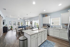 A spacious kitchen with white cabinets, a central island, and a dining area with a blue rug. - New Coastal Retreat | Boardwalk, Fire Pit & Games (Port St. Joe)