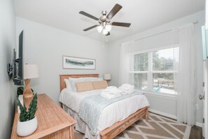 A bedroom with a bed, a ceiling fan, a window with blinds, a bedside table with a lamp, and a potted plant. - New Coastal Retreat | Boardwalk, Fire Pit & Games (Port St. Joe)