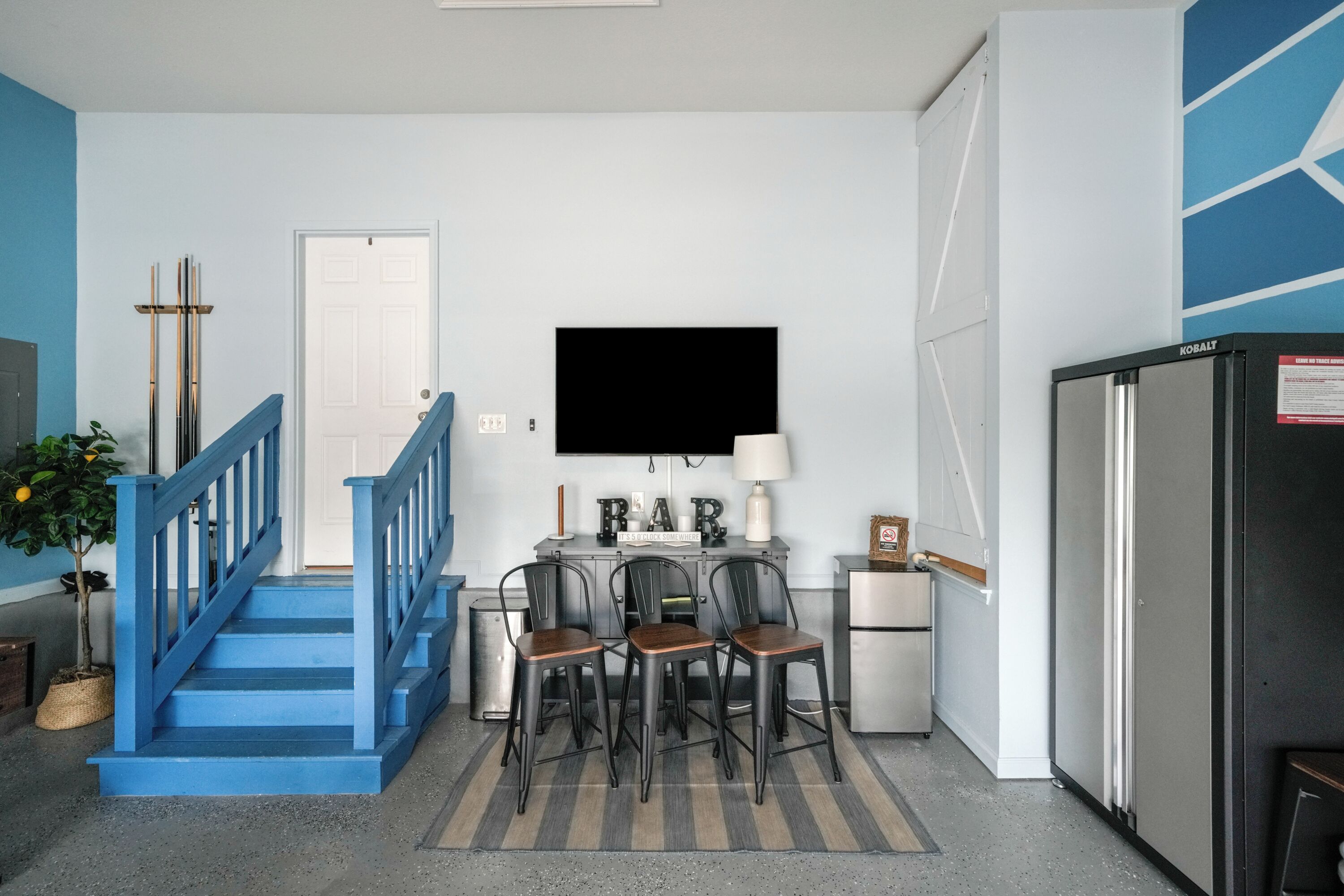 A kitchen with a refrigerator, a TV mounted above a counter, and a staircase with blue railings leading to another room.