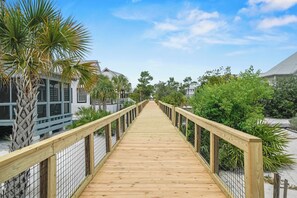 A wooden boardwalk leading to a building with a white facade and dark windows. - New Coastal Retreat | Boardwalk, Fire Pit & Games (Port St. Joe)