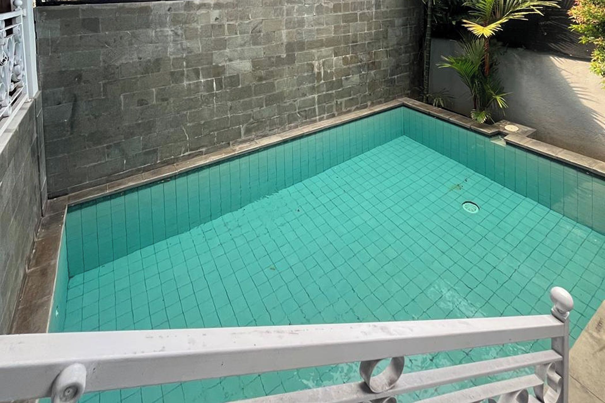 Una piscina pequeña y rectangular con suelo de baldosas, rodeada por un muro de piedra y una barandilla blanca.