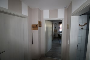 Un couloir avec une porte à gauche, une étagère en bois et un mur recouvert de carreaux à droite.