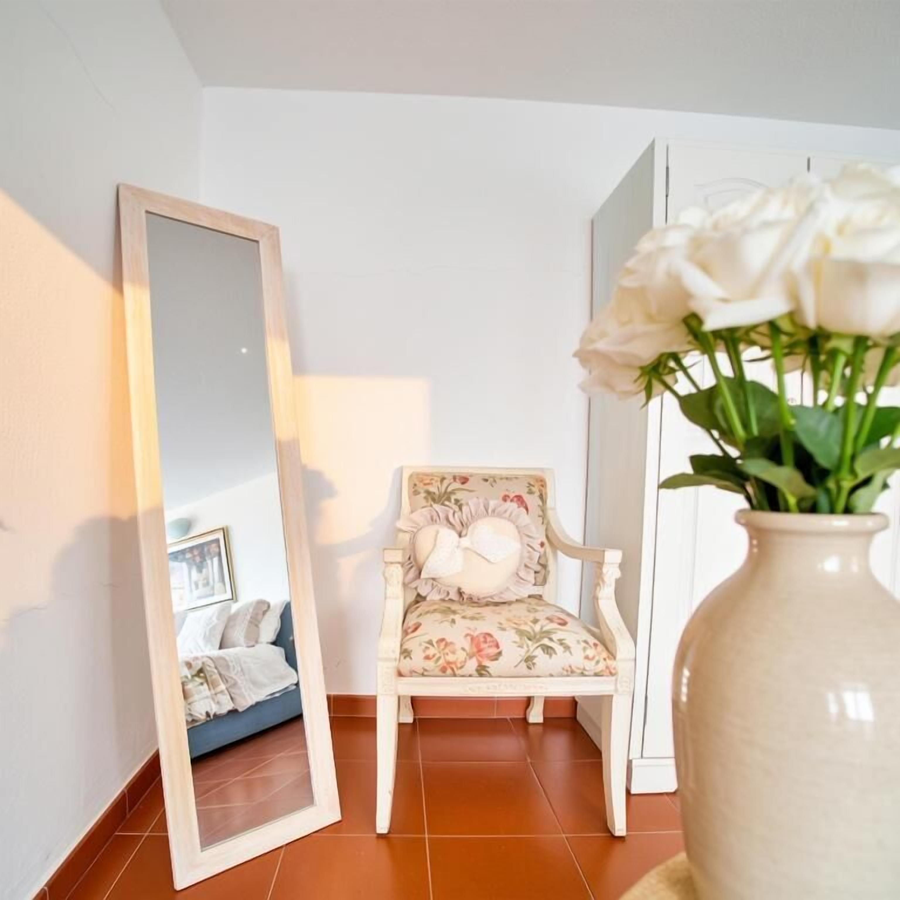 Una habitación con un espejo grande, un sillón estampado con motivos florales y un florero con flores.