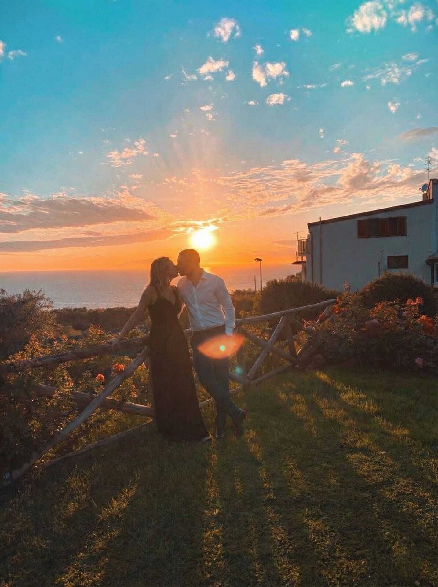 A couple standing in a grassy area with a sunset in the background.