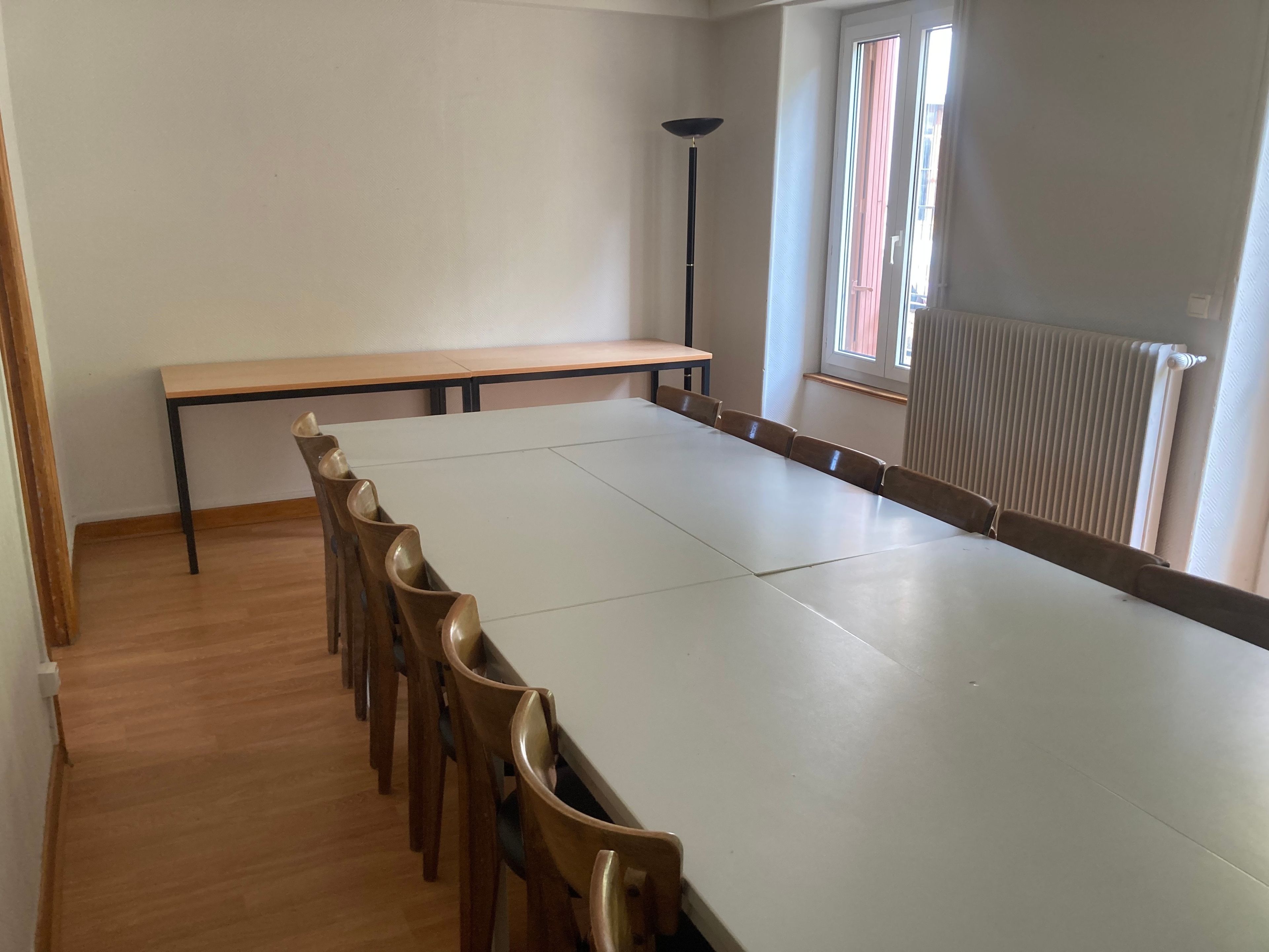 Een vergaderruimte met een lange tafel, stoelen, een staande lamp en een raam met zonwering.