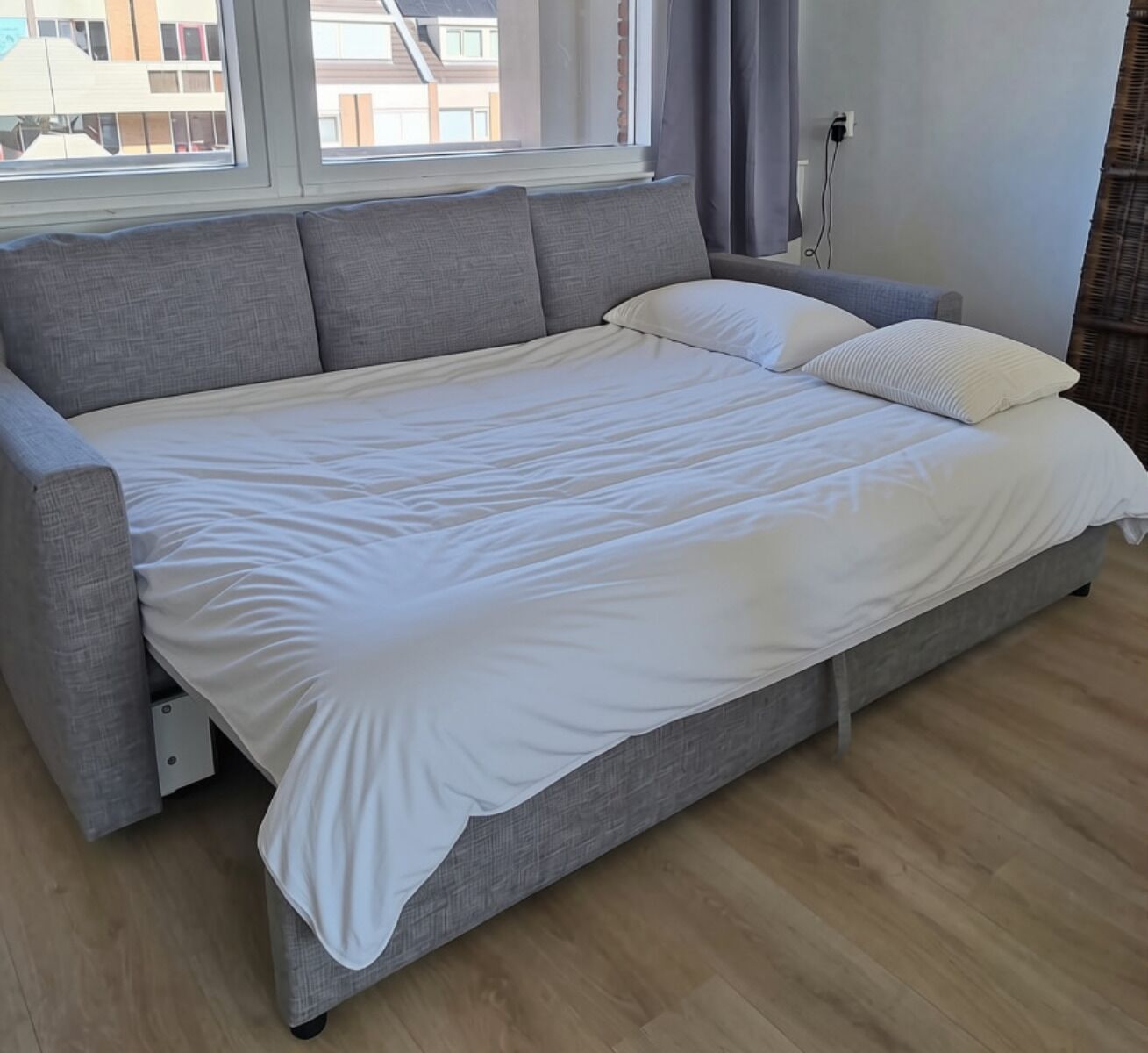 A modern bedroom with a grey sofa bed, white bedding, a window with curtains, and a visible power outlet.