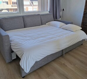 A modern bedroom with a grey sofa bed, white bedding, a window with curtains, and a visible power outlet. - The beach, the ocean, and the dunes right outside your door! This Duinparel apartment is located on the 2nd floor of the Kennedyflat complex. This apartment is on the ground floor and features one bedroom, a kitchen, a bathroom, and a west-facing balcony. (Egmond aan Zee)