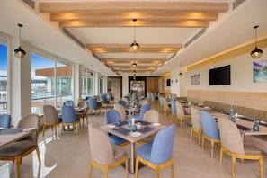 A spacious dining area with multiple tables and chairs, large windows offering a view, and a high ceiling with wooden beams.
