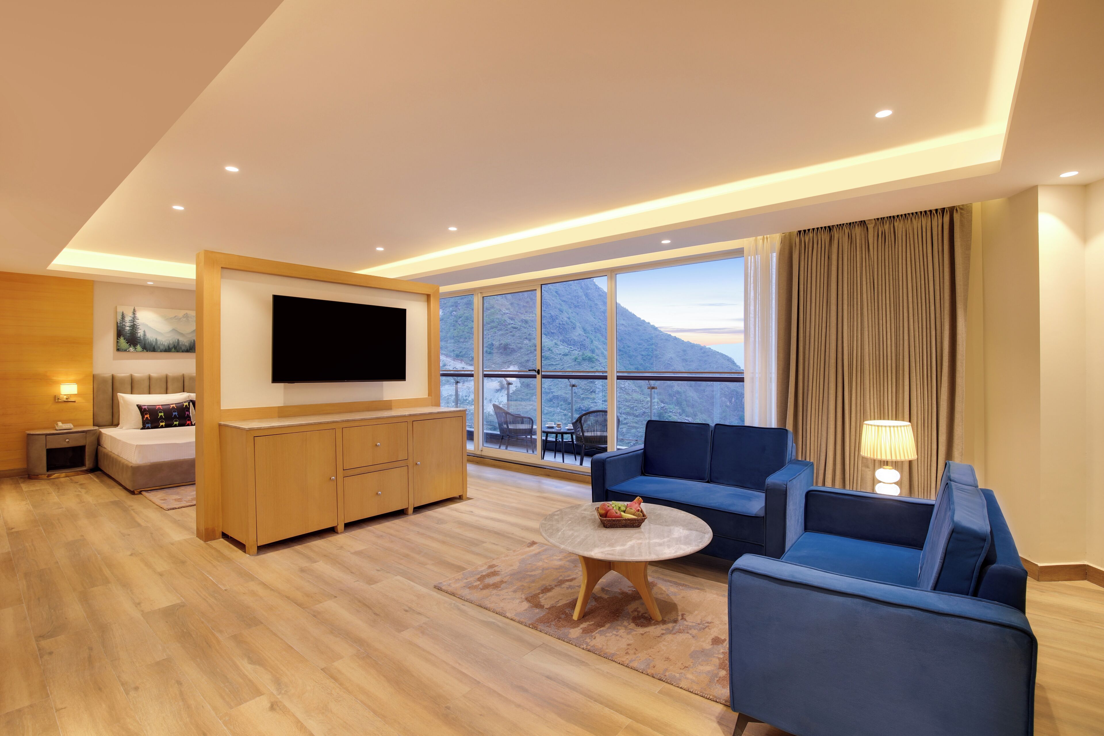 Habitación de hotel moderna con televisor de pantalla plana, sofá, mesita de café y un amplio ventanal con vistas a la montaña.