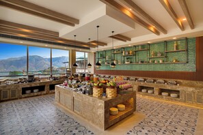 Zona de desayuno de hotel con bufé, vistas a montañas y un salpicadero de azulejos verdes.