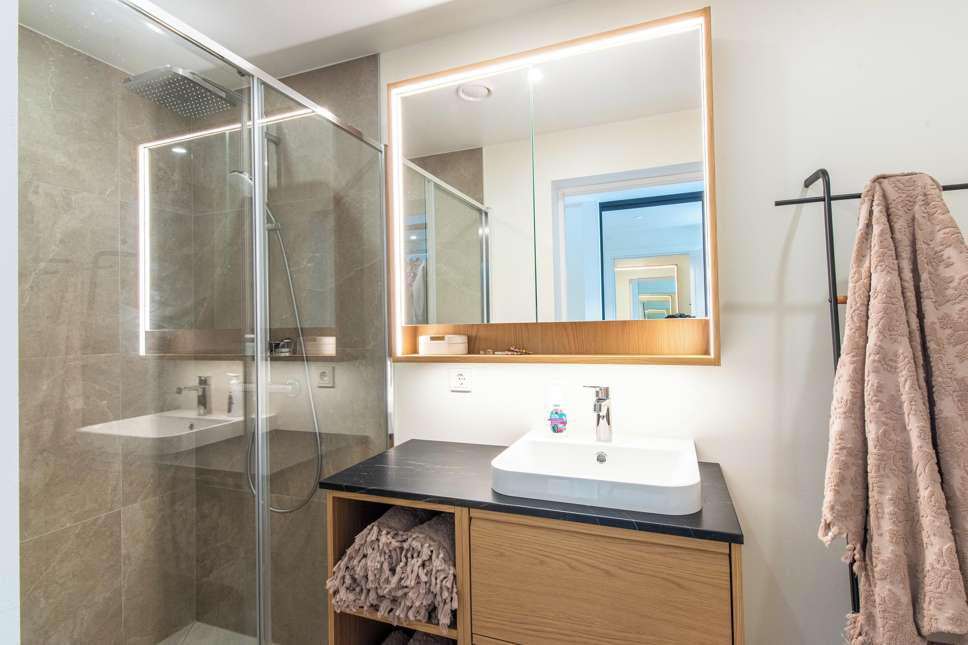 Een moderne badkamer met een douchecabine van glas, een witte wastafel met houten opbergruimte, en een grote spiegel erboven.