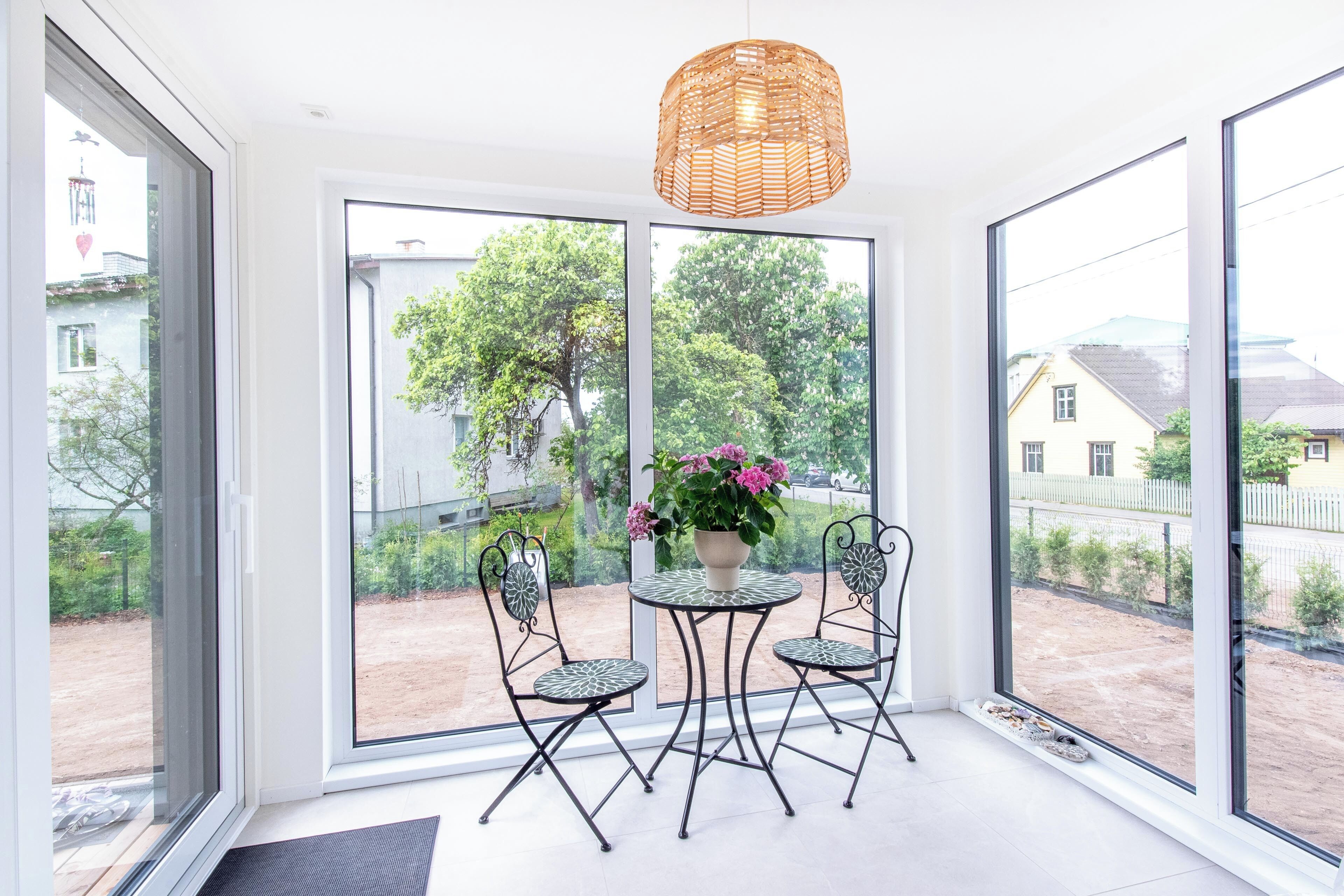 Een kamer met een groot raam, een rieten hanglamp, een ronde tafel met stoelen en een bloeiende plant in een pot.