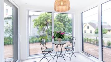 Een kamer met een groot raam, een rieten hanglamp, een ronde tafel met stoelen en een bloeiende plant in een pot.
