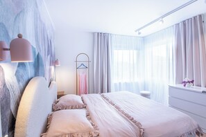 A bedroom with a bed, a bedside table, a hanging light fixture, a window with curtains, and a wall-mounted towel rack.