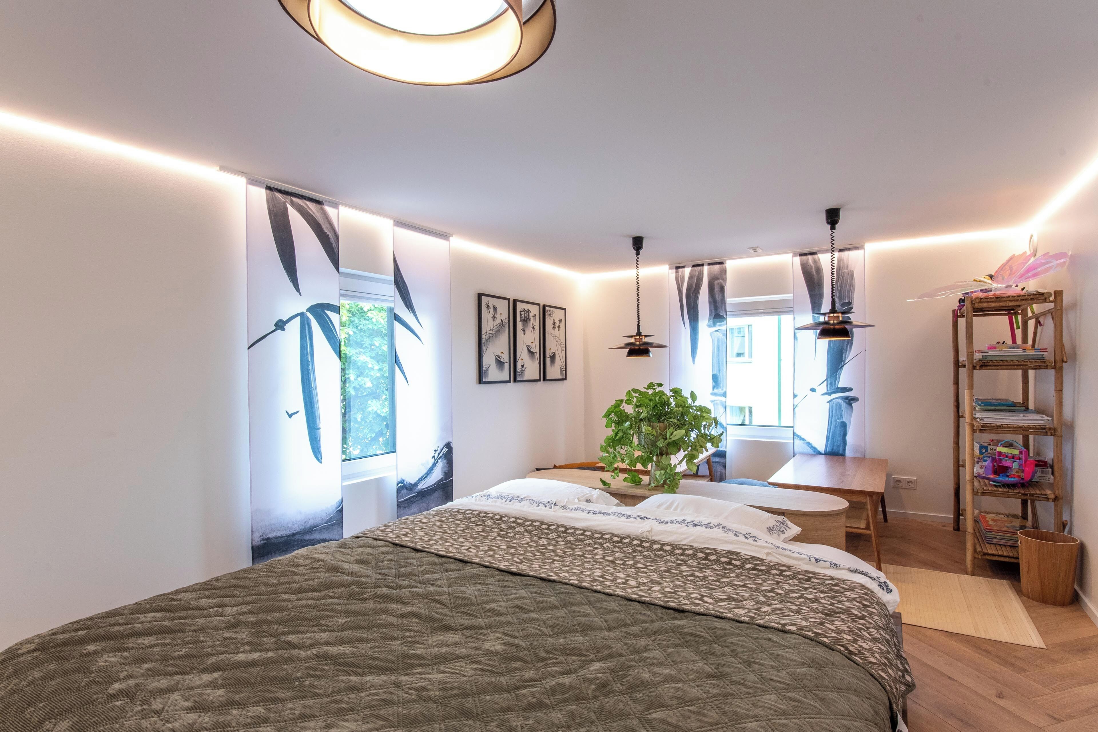 Ein modernes Schlafzimmer mit einem großen Bett, einem Holfnachtkästchen, einer hängenden Pflanze und einem Bücherregal.