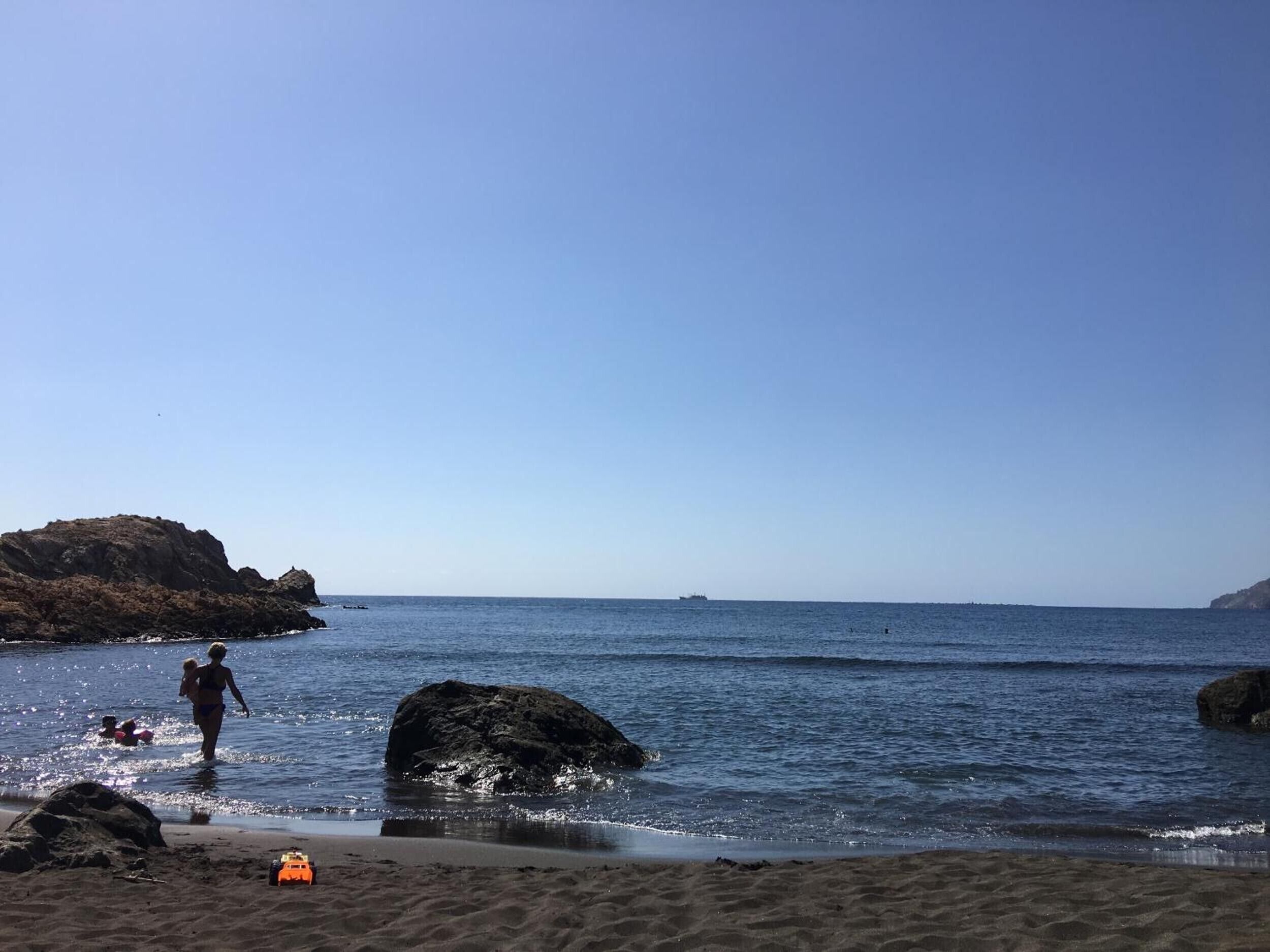 Una playa con rocas, cielo despejado y un barco a lo lejos.