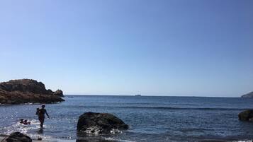 Una playa con rocas, cielo despejado y un barco a lo lejos.