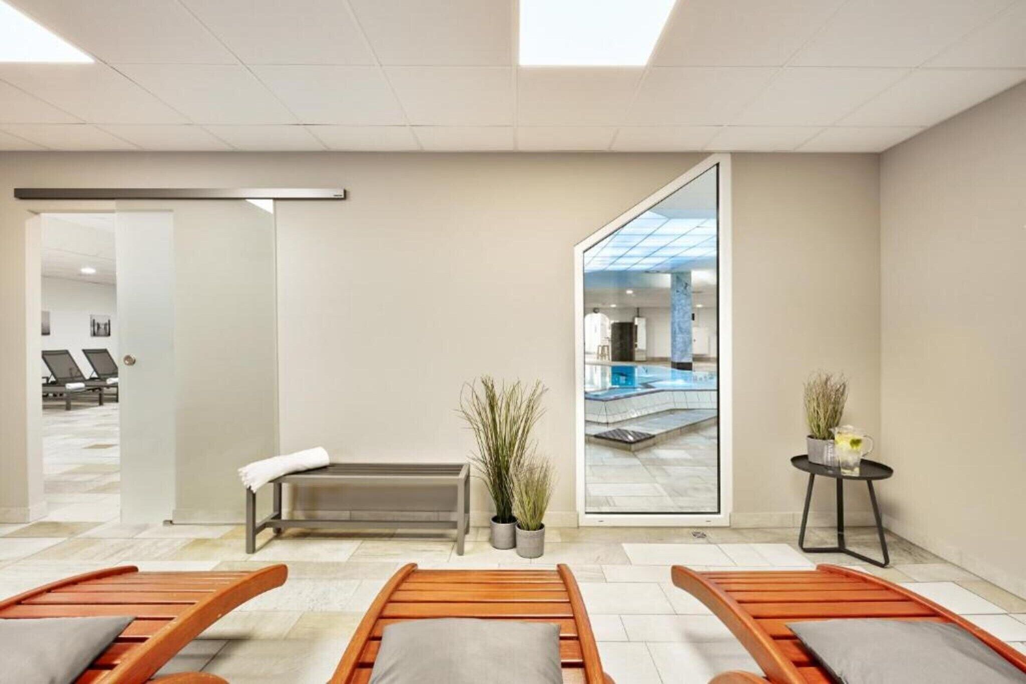 Ett modernt spa-rum med en bänk, två solstolar, ett litet bord med en växt och en glasdörr som leder ut till poolområdet.