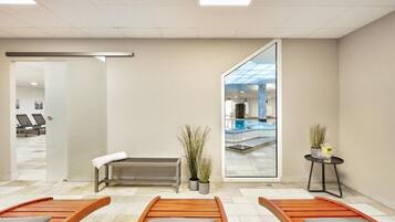 Ett modernt spa-rum med en bänk, två solstolar, ett litet bord med en växt och en glasdörr som leder ut till poolområdet.