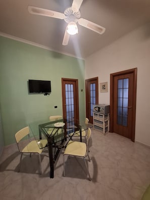 Un comedor con una mesa de vidrio y sillas, un ventilador de techo y un televisor montado en la pared.