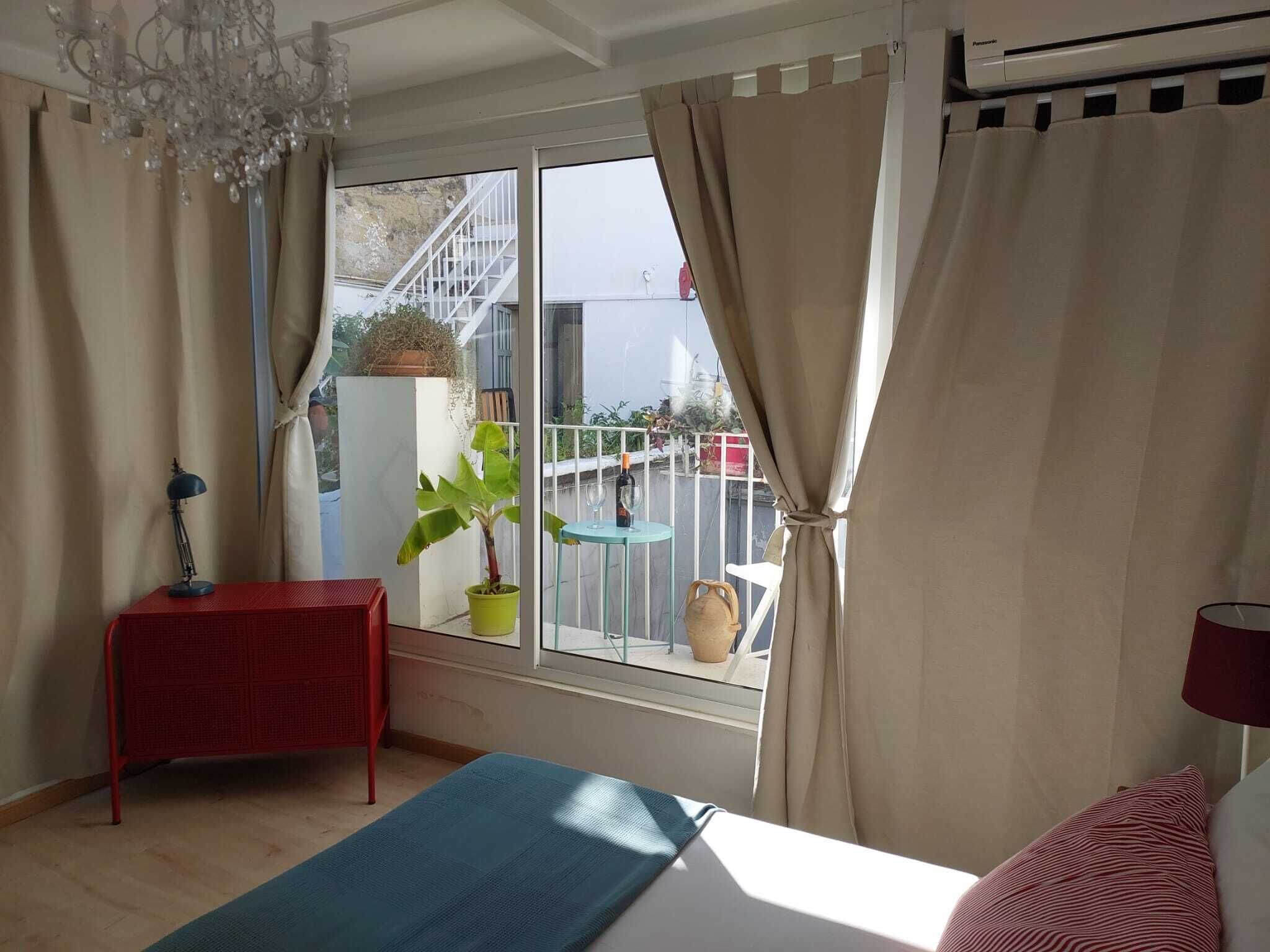 Ein Schlafzimmer mit einem Bett, einem roten Nachttisch, einer Topfpflanze und Blick auf einen Balkon mit Gartenmöbeln.