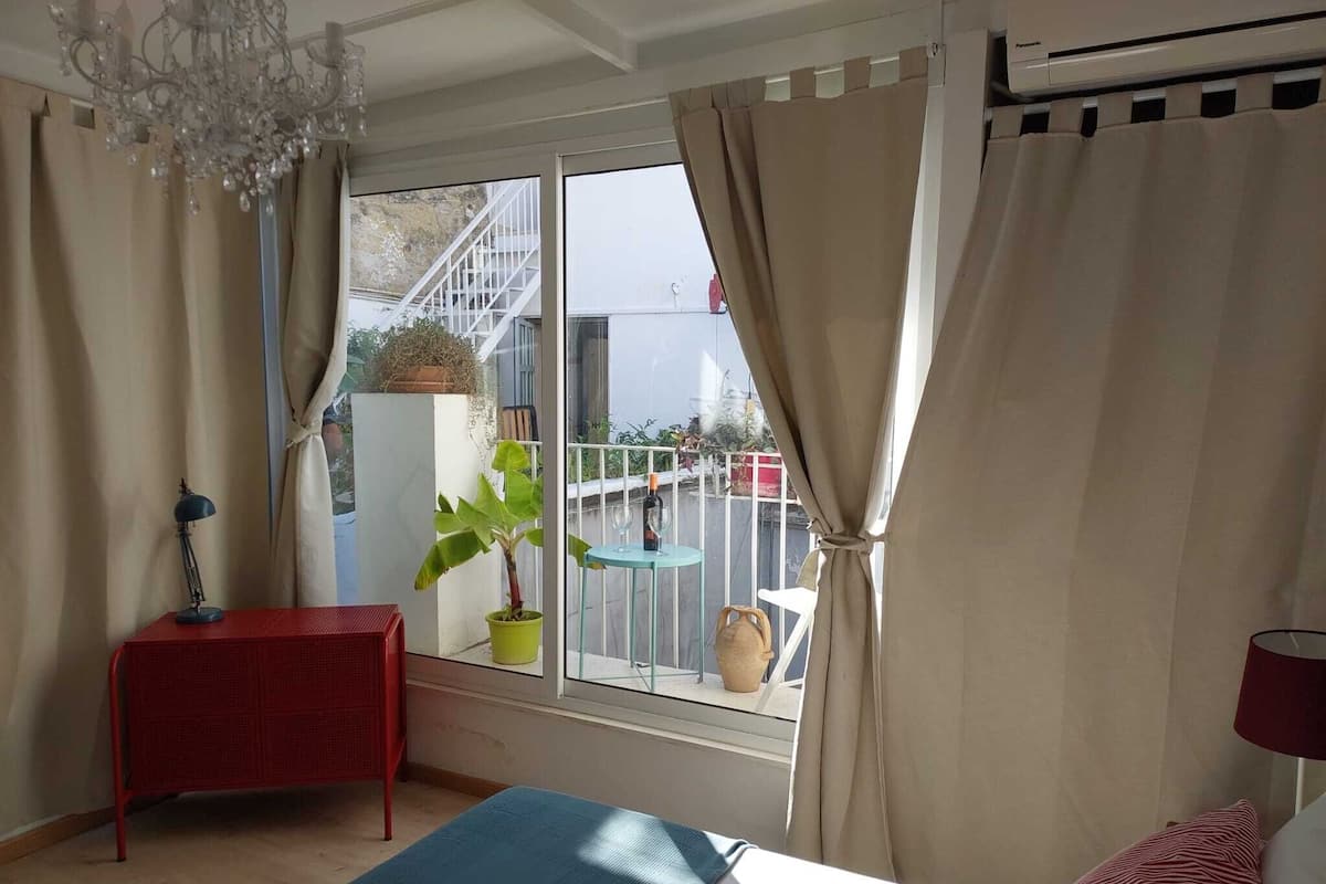Un dormitorio con cama, una mesita de noche roja, una planta en maceta y vistas a un balcón con mobiliario exterior.