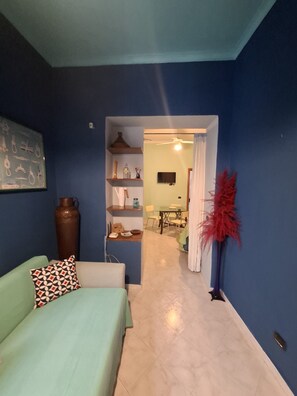 Una habitación con pared azul, un sofá verde con cojines estampados, un estante de madera con diversos objetos y un área de comedor al fondo.
